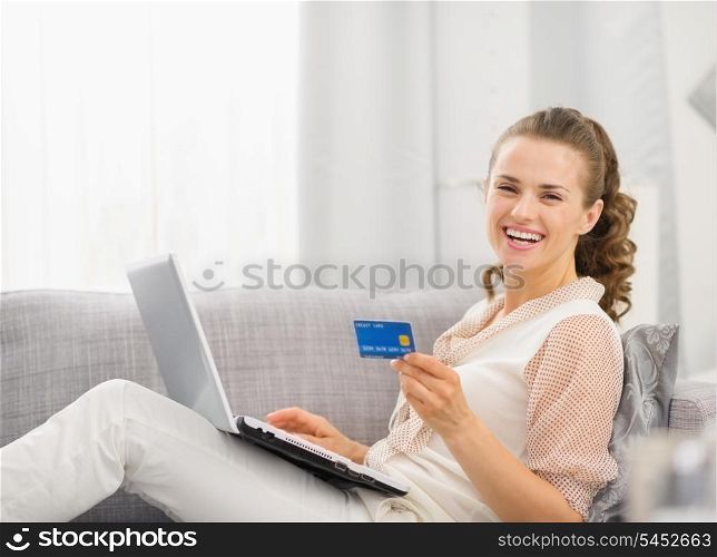 Happy young housewife laying on sofa with laptop and credit card