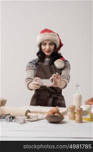 Happy young housewife making christmas cookies in kitchen and taking pictures on smartphone