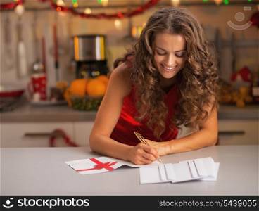 Happy young housewife signing christmas postcards in kitchen