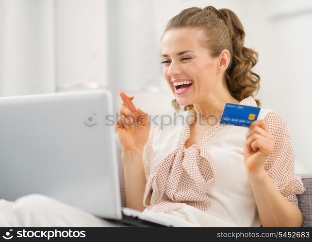 Happy young housewife with laptop and credit card crossed fingers