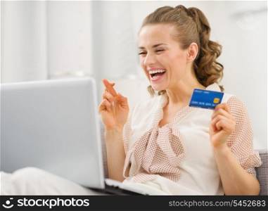 Happy young housewife with laptop and credit card crossed fingers