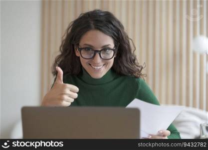 Happy young Italian female employee in glasses talks online on laptop with colleagues or business partner, showing thumb up, cheerful Spanish woman freelancer making video call while working remotely. Happy young Italian female employee talks online on laptop with colleagues and showing thumb up