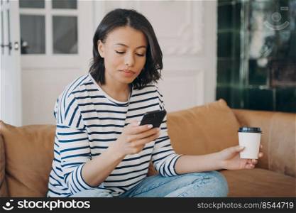 Happy young lady is texting on telephone. European woman having rest alone with cup of coffee while sitting on leather couch. Leisure and relaxation at home. Communication technology.. Happy young lady is texting on telephone. European woman having rest with cup of coffee at home.
