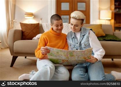 Happy young lesbian woman couple looking at paper map planning trip together while relaxing in living room at home. Happy young lesbian woman couple looking at paper map planning trip
