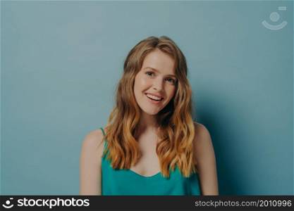 Happy young light brown-haired woman posing with head tilted, looking at camera with gentle smile opening her mouth slightly shows perfectly straight teeth isolated over blue studio background. Happy young woman posing isolated over blue studio background, looking at camera with gentle smile