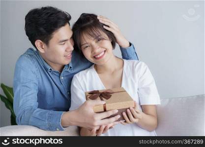 Happy young love asian couple sitting on a couch at home looking at a gift box brown
