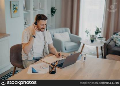 Happy young man adjusting headset while studying online on laptop or having web conference during remote work at home, male freelancer having video call and making some notes, sitting at his workplace. Happy male freelancer talking online and making notes while sitting at his workplace at home