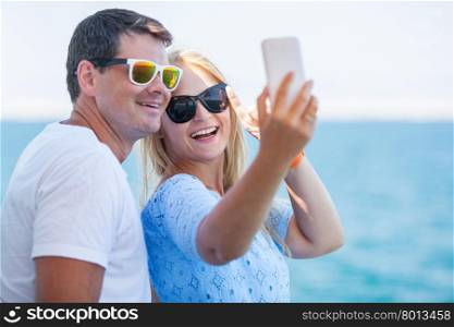 Happy young man and woman in fashionable sunglasses taking cellphone selfie on background of defocused blue sea. Vacation photos. Happy summer selfie of young couple in sunglasses