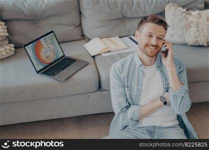 Happy young man calling family to inform about receiving promotion at his company after submitting successful project idea, sitting on floor while resting against couch with open laptop showing graphs. Happy young man calling family to inform about receiving promotion