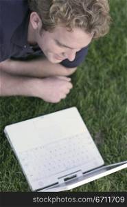 Happy young man dressed in blue shirt relaxes and lies down in the green grass, looking at his white laptop