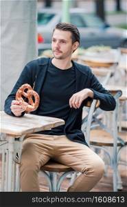 Happy young man eating pretzel outdoors in Europe. Beautiful young man holding pretzel and relaxing in park