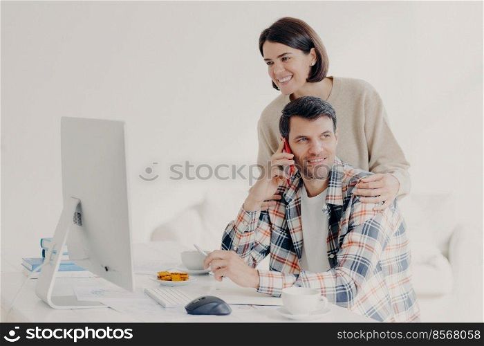 Happy young man holds mobile phone, discusses issues, manages home accounts with wife, pose in coworking space, sit in front of computer, discuss family expenses. Couple collaborate together