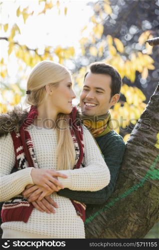 Happy young man hugging woman while leaning on tree trunk during autumn in park