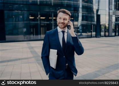 Happy young man in blue suit with newspaper and hand in pocket calling his wife to tell her about promotion, standing while radiating confidence and success in front of glass office building. Happy office worker with newspaper talking on smartphone