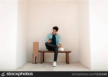 Happy Young Man in Casual wear Using Mobile Phone while Sitting on Bench by the Wall. Lifestyle of Modern People. Wide Shot, Full Length