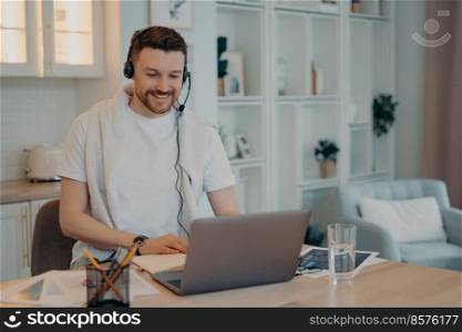 Happy young man in white shirt wearing headphones sitting at his workplace in living room and using laptop during online call or internet lesson. Online study and freelance concept. Male freelancer looking at laptop screen while working at home