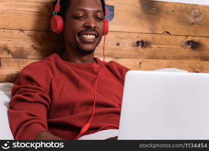 Happy young man listening music with headphones and using laptop on his bed. Indoors