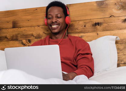 Happy young man listening music with headphones and using laptop on his bed. Indoors