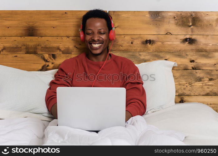 Happy young man listening music with headphones and using laptop on his bed. Indoors