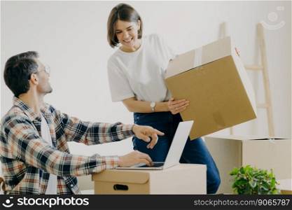 Happy young man points into laptop computer, searches new design for flat, busy woman carries boxes, makes purchase on new habitation, search home decorations, connected to wirless internet.