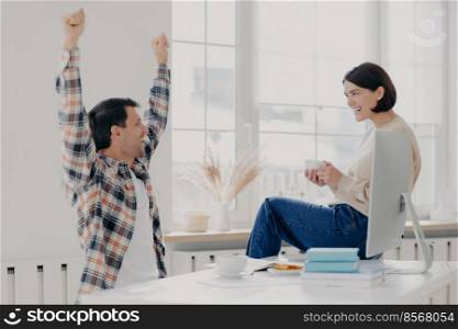 Happy young man rejoices successfully finished work, raises arms, dressed in checkered shirt, being glad, shares his success with wife who sits on desktop with coffee cup, collaborate together