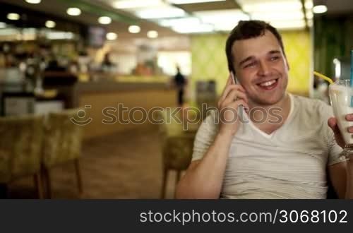 Happy young man sitting chatting on his mobile in a club , hotel or pub with a glass of tropical cocktail in his hand