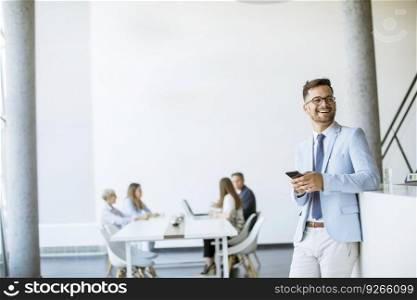 Happy young man using his mobile phone in the office and smiling while his colleagues working in the background