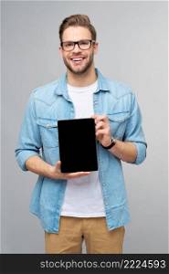 Happy young man wearing jeans shirt standing and holding tablet pc pad over studio grey background.. Happy young man wearing jeans shirt standing and holding tablet pc pad over studio grey background