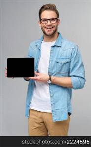 Happy young man wearing jeans shirt standing and holding tablet pc pad over studio grey background.. Happy young man wearing jeans shirt standing and holding tablet pc pad over studio grey background