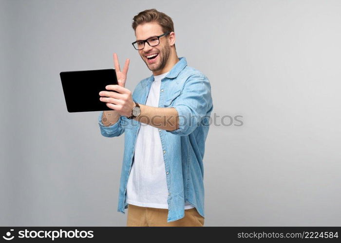 Happy young man wearing jeans shirt standing and using tablet over studio grey background.. Happy young man wearing jeans shirt standing and using tablet over studio grey background