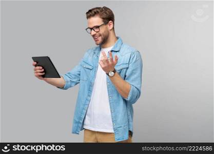 Happy young man wearing jeans shirt standing and using tablet over studio grey background.. Happy young man wearing jeans shirt standing and using tablet over studio grey background