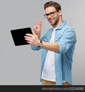 Happy young man wearing jeans shirt standing and using tablet over studio grey background.. Happy young man wearing jeans shirt standing and using tablet over studio grey background