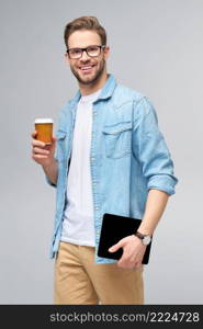 Happy young man wearing jeans shirt standing and using tablet pc pad and holding cup of coffee to go standing over studio grey background.. Happy young man wearing jeans shirt standing and using tablet pc pad and holding cup of coffee to go standing over studio grey background