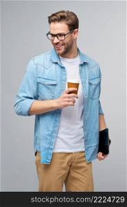 Happy young man wearing jeans shirt standing and using tablet pc pad and holding cup of coffee to go standing over studio grey background.. Happy young man wearing jeans shirt standing and using tablet pc pad and holding cup of coffee to go standing over studio grey background