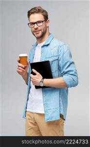 Happy young man wearing jeans shirt standing and using tablet pc pad and holding cup of coffee to go standing over studio grey background.. Happy young man wearing jeans shirt standing and using tablet pc pad and holding cup of coffee to go standing over studio grey background