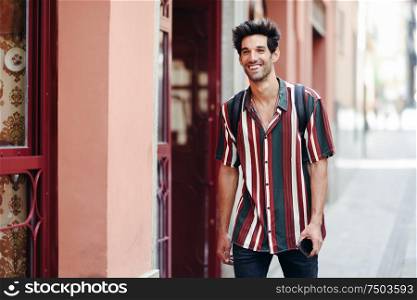 Happy young man with dark hair and modern hairstyle wearing casual clothes in urban background.. Attractive young man with dark hair and modern hairstyle wearing casual clothes outdoors