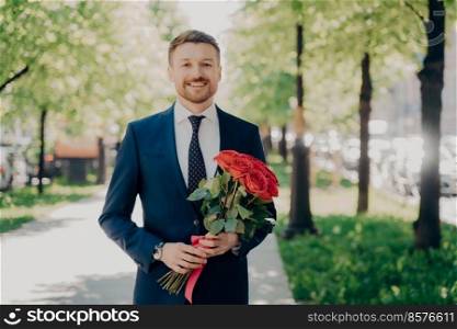 Happy young man with neat beard in elegant formal dark blue suit walking joyfully along green alley in park with beautiful bouquet of red roses in his hands in sunny day to meet his lovely date. Happy young man in elegant suit with bouquet of flowers walking in park