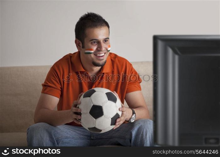 Happy young man with painted face watching television while holding soccer ball