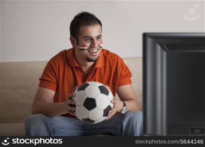 Happy young man with painted face watching television while holding soccer ball