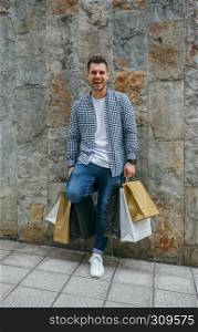 Happy young man with shopping bags leaning on a stone wall. Young man with shopping bags