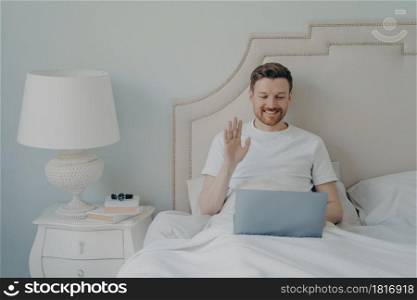Happy young man with stubble waving hand to laptop while having online video call and sitting in bed, making conversation with his close friends during leisure time on weekend at home. Relaxed young man with stubble using laptop laptop while sitting in bed