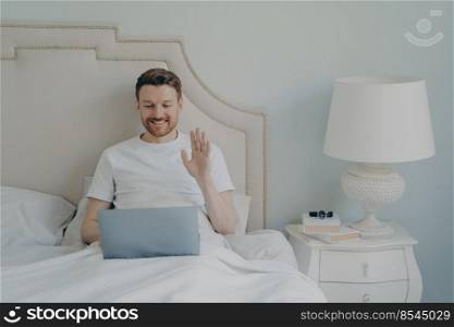 Happy young man with stubble waving hand to laptop while having online video call and sitting in bed, making conversation with his close friends during leisure time on weekend at home. Relaxed young man with stubble using laptop laptop while sitting in bed