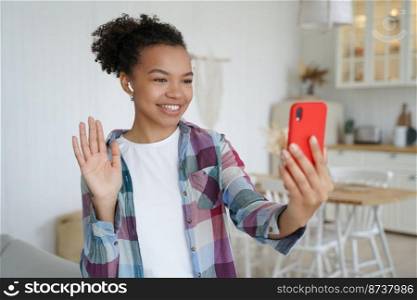 Happy young mixed race girl answering video call, holding phone glad to meet friend online, waving hand to smartphone camera, saying hello. Smiling teen lady chatting makes greeting gesture at home.. Smiling mixed race girl holds phone, makes greeting gesture, talking online on video call at home