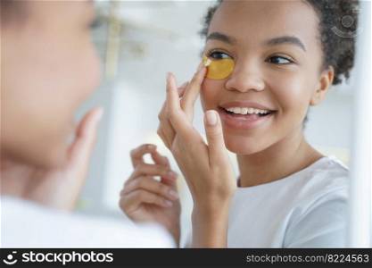 Happy young mixed race girl applies under eye patches to moisturize facial skin, reduce dark circles, remove fine wrinkles, looking in mirror. Beauty treatment, skin care morning routine concept.. Young mixed race girl applies under eye patches for face skin, looking in mirror. Skincare treatment
