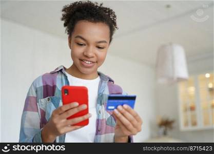 Happy young mixed race girl holding credit card and smartphone, transferring money, shopping in internet store. Modern teen lady using mobile bank app, online banking services, standing at home.. Happy teen mixed race girl holding credit card, phone, uses mobile bank app, online banking services