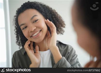 Happy young mixed race girl looks at mirror, touching face after applying moisturizing mask or cream. Smiling pretty biracial lady enjoying smooth healthy facial skin. Natural beauty, skincare concept. Happy young mixed race girl looks at mirror, enjoying healthy facial skin. Natural beauty, skincare