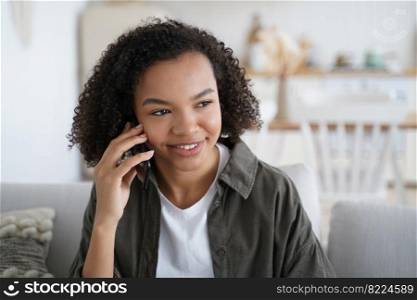 Happy young mixed race girl making phone call at home, friendly smiling teen lady chatting, enjoying pleasant mobile conversation, holding smartphone while talking, sitting in living room.. Friendly smiling mixed race teen girl makes phone call chatting enjoying mobile conversation at home