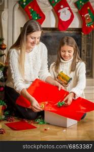 Happy young mother and cute daughter packing Christmas gifts in red paper and tying with golden ribbon