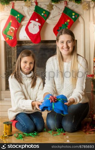 Happy young mother and daughter sitting on floor at fireplace and packing sweater for Christmas present