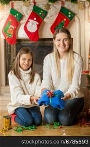 Happy young mother and daughter sitting on floor at fireplace and packing sweater for Christmas present
